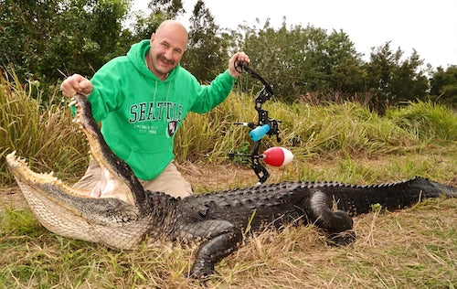 Man in a green sweatshirt holding a bow and arrow next to a large alligator he hunted in a grassy area with Florida Bowfishing Charters.