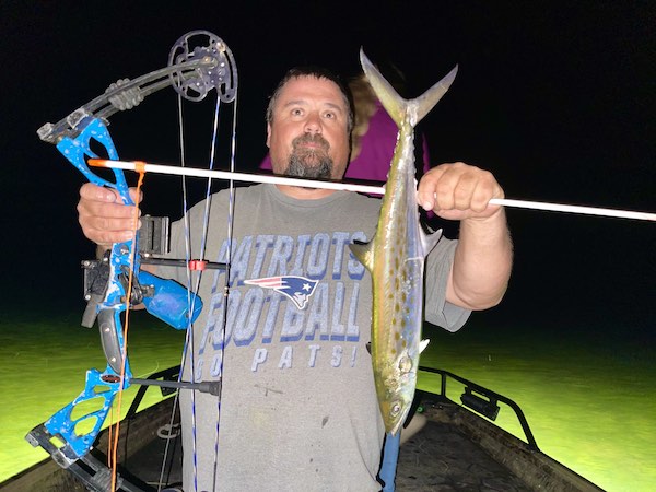 Man holding a bow and arrow with a fish he has caught at night while bowfishing.