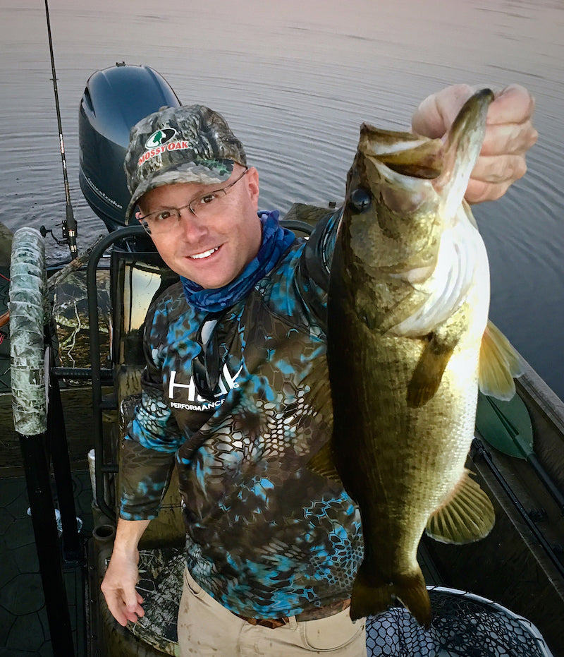 Man holding a large bass on a fishing charter boat, wearing a cap and glasses.
