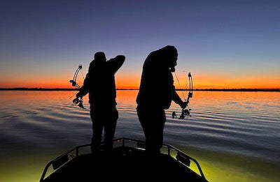 Two people bowfishing from a boat at sunset on a lake