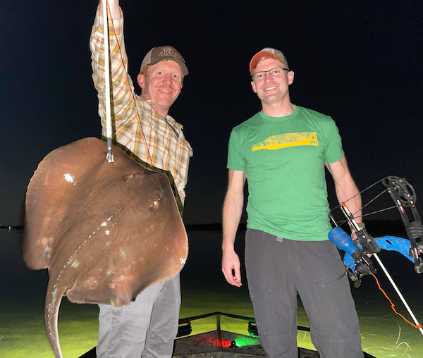 Two men standing next to a large stingray caught with a bow and arrow at night.