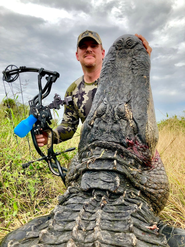 Man holding a large alligator with a bow and arrow in a grassy area an a Florida Gator Hunt.