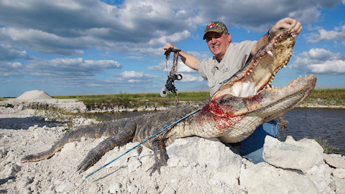Man holding a large alligator with a bow and arrow, standing on a rocky shore with a cloudy sky.